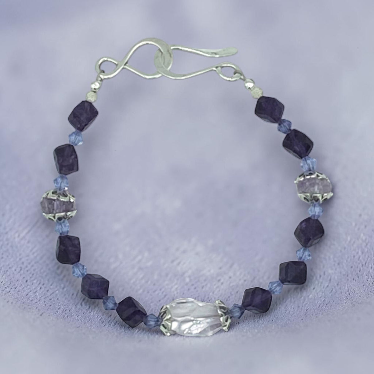 A beaded bracelet featuring dark purple faceted beads, light blue faceted beads, a large textured clear crystal centerpiece, and silver-toned metal spacers and clasp.