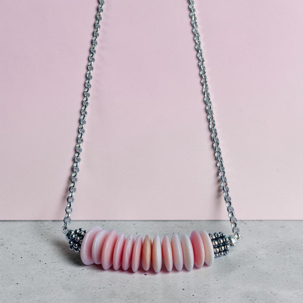 A silver chain necklace featuring a horizontal bar of small, disc-shaped beads in shades of pink and white, displayed against a split pink and gray background.