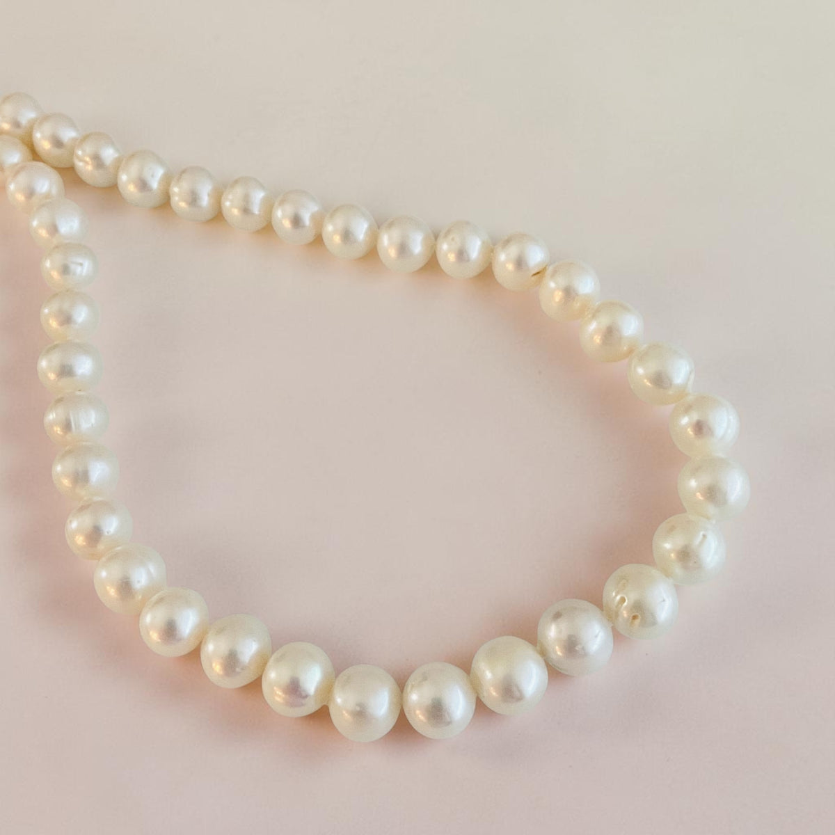 A strand of cream freshwater pearls arranged in a loose circle on a soft, pale pink background.