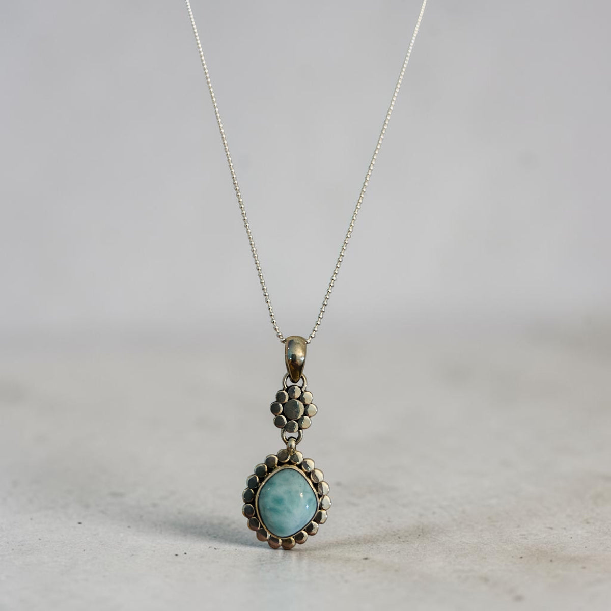 A silver-toned dangle pendant necklace featuring a light blue-green marbled stone in a scalloped metal frame, suspended from a delicate chain against a neutral gray background.