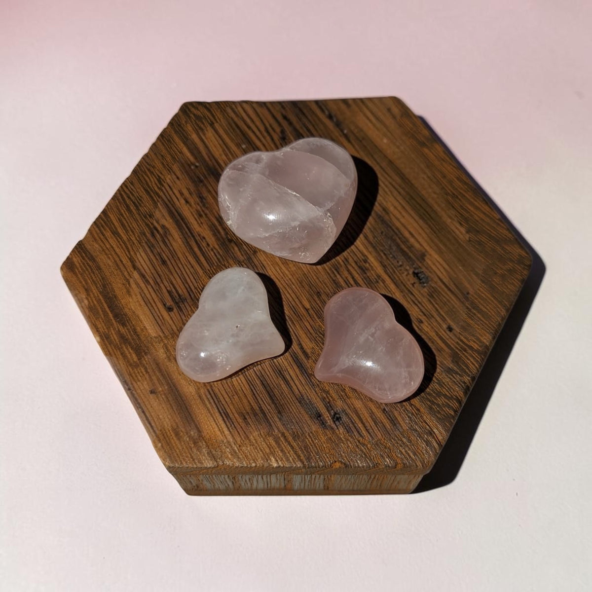 Three heart-shaped rose quartz crystal points resting on a hexagonal wooden block against a light pink background.
